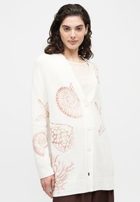 Νεαρή γυναίκα που φοράει λευκή ζακέτα με κουμπιά και μοτίβα από κοχύλια και κοράλλια, στέκεται μπροστά σε ένα απλό ανοιχτό φόντο.