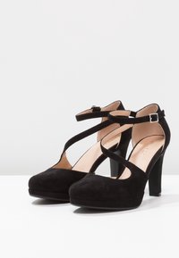 Chaussures à talons hauts en daim noir avec une plateforme, dotées de brides croisées autour de la cheville et d'un design ouvert sur le côté. Texture lisse et talon robuste.