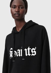 Felpa nera in cotone, con una grande grafica bianca "Saints". Include un cappuccio con coulisse e una leggera texture. Etichetta di cura visibile.