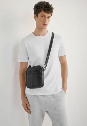 Młody mężczyzna w białej koszulce i szarych spodniach, niosący czarną skórzaną torbę na ramię z zamkami błyskawicznymi.