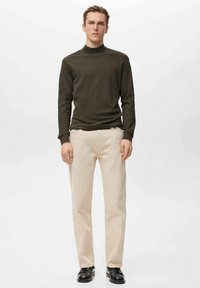 Olivgrüner Rollkragenpullover, leichter Stoff, schlanke Passform; kombiniert mit cremefarbenen Hosen, gerade geschnitten, getragen mit schwarzen Schuhen.