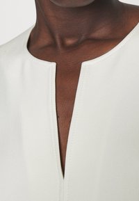 Tissu blanc et doux avec un décolleté en V. Présente des coutures soignées le long de l'encolure, une texture lisse et une vue rapprochée du vêtement.