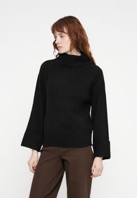 Pull en maille noire avec un col haut, des manches larges et des poignets côtelés, accompagné d'un pantalon marron. Texture lisse, design décontracté.