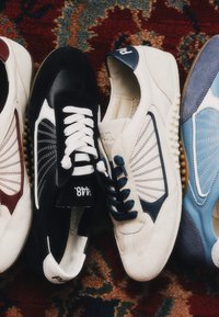 Quatre paires de baskets style rétro en blanc, noir, crème et bleu placées côte à côte sur un tapis à motifs.