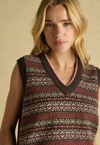 Blondinka v brezrokavnem pletenem jumperskem telovniku z rdečim, zelenim, belim in rjavim Fair Isle vzorcem na bež ozadju.