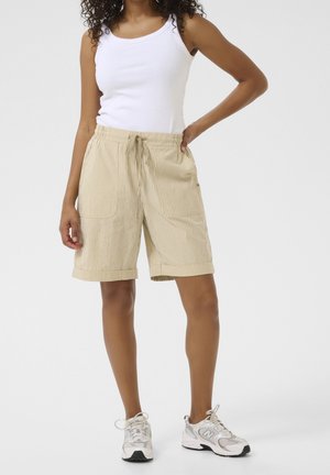 Femme portant un short beige rayé longueur genou avec cordon de serrage et un débardeur blanc, debout avec une main sur la hanche et des baskets blanches.
