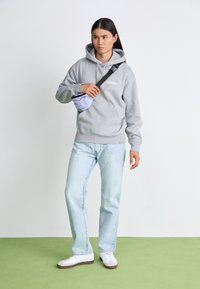 Grå hoodie med framficka, "Dickies"-logotyp. Ljusblå jeans med rak ben. Modellen håller en ljuslila midjeväska och har på sig vita sneakers.