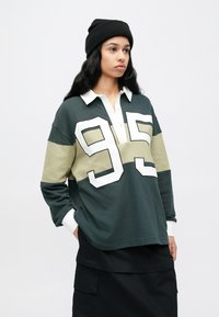 Πράσινο και μπεζ μακρυμάνικο πόλο με λευκό γιακά και μεγάλο λευκό αριθμό "95". Φορεμένο με μα�ύρο σκουφάκι και φούστα. Υφή υφάσματος.