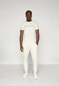 Krem T-skjorte med "BJÖRN BORG" tekst, parret med matchende krem joggebukser. Begge er laget av myk stoff; modellen har på seg hvite joggesko.