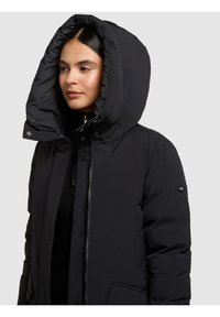 Sort polstret jakke med stor, struktureret hætte, lynlåslukning med trykknapper og sidelommer. Glat tekstur og minimalistisk design.