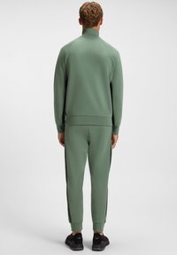 Žalios spalvos sportinis kostiumas su aukštu apykakliu, ribinėmis rankogaliais ir šoninėmis juostomis. Pagamintas iš minkšto audinio su lygiu paviršiumi. Juodi sportbačiai.