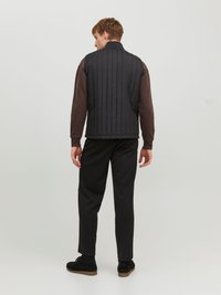 Gilet matelassé noir avec un col montant, doté de coutures verticales. Porté par-dessus une chemise à manches longues marron foncé et un pantalon noir.