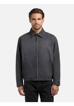 Mężczyzna ubrany w ciemnoszarą kurtkę zapinaną na zamek z kołnierzem i przednimi kieszeniami, stojący na tle jasnego, jednolitego tła.