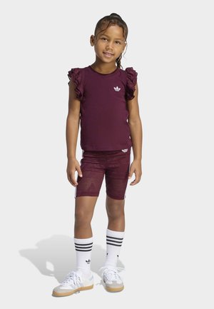 Ung pige stående, iført en bordeauxbluse med flæser på ærmerne, matchende shorts, hvide sokker med striber og hvide sneakers på en lys baggrund.