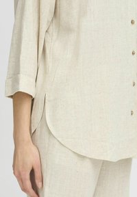 Camicia in lino beige con vestibilità rilassata, maniche a tre quarti, orlo arrotondato e chiusura con bottoni. Texture liscia con un delicato dettaglio intrecciato.