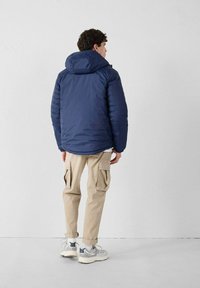 Hombre con chaqueta acolchada azul marino con capucha, pantalones cargo beige y zapatillas blancas, de pie frente a una pared blanca con las manos relajadas a los lados.