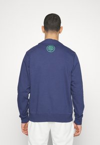 Felpa blu navy con collo rotondo e maniche lunghe. Presenta un logo verde circolare sulla parte superiore della schiena. Tessuto morbido e strutturato.