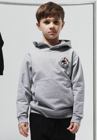 Sudadera gris con bolsillo frontal, que cuenta con un parche de logo de un bulldog. Tela suave, corte relajado y puños acanalados. Llevada sobre pantalones negros.