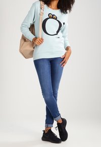 Vrouw in een lichtblauwe sweatshirt met een cartoonogenontwerp, blauwe opgerolde jeans, zwarte sneakers en een beige rugzak over één schouder.