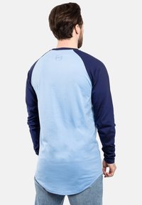 Svijetloplata majica dugih rukava s mornarskim raglan rukavima. Mekani materijal, zaobljen rub i izvezeni logo na stražnjem izrezu vrata.