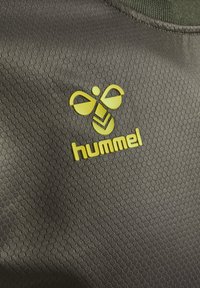 Hummel-logotyp på mörkgrå strukturerad tyg, med ett distinkt bikake-mönster och en starkt gul emblem som visar varumärkets namn och design.