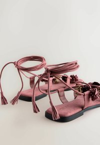 Ružové semišové ploché sandále s čiernymi podrážkami, viazaním okolo členkov a dekoratívnymi kvetinovými ozdobami na remienkoch.