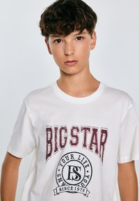 Valge puuvillane T-särk, millel on suur burgundia "BIG STAR" graafika ja ümmargune logo, millel on must tekst "YOUR LIFE SINCE 1979".