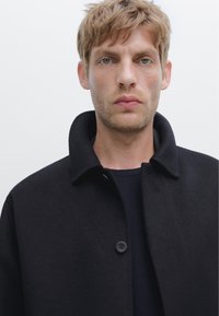 Cappotto in lana nera con colletto strutturato, chiusura con un solo bottone e superficie liscia. Indossato sopra una camicia blu navy, design minimalista.