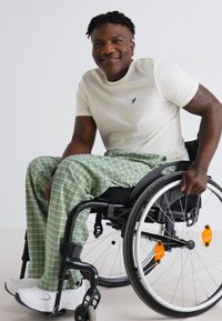 Fehér póló és zöld kockás nadrág egy kerekesszékben ülő személynél. A kerekesszék fekete és narancssárga díszítésekkel rendelkezik, a kerekeken küllővédők találhatók.