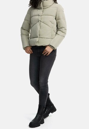 Femme portant une veste d'hiver matelassée beige clair, un jean skinny noir et des bottines noires épaisses, debout avec les mains près de la taille.