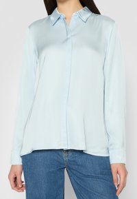 Camicia azzurro chiaro a maniche lunghe in satin. Presenta un colletto, chiusura con bottoni e una vestibilità rilassata con spacco laterale all'orlo.