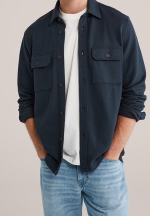 Mann trägt marineblaue Hemdjacke mit Knöpfen über weißem T-Shirt und hellblauen Jeans mit Händen in den Taschen.