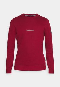 Sweatshirt rouge à manches longues, col rond et poignets côtelés. Présente le texte "REDEFINED REBEL" sur le devant. Texture lisse, design décontracté.