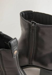 Bottes chevilles en cuir noir avec fermeture éclair latérale et languette, présentant une texture lisse et des talons blocs bas, affichées sur un fond uni.