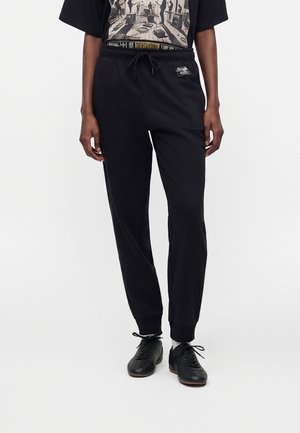 Personne portant un pantalon de jogging noir avec taille à cordon et des chaussures noires à lacets, debout sur un fond blanc.