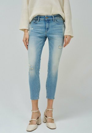 Lichtblauwe denim jeans met versleten details, taps toelopende pasvorm en verkorte lengte. Draagt een beige block-heel schoenen en een crèmekleurige trui.