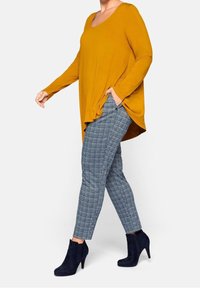 Senapsgul V-ringad långärmad topp i kombination med figurnära rutiga byxor i blå nyanser och marinblå ankelboots med klack.