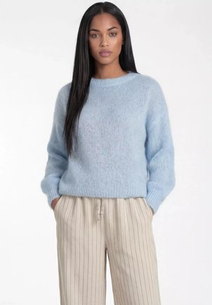 Kobieta z długimi czarnymi włosami, ubrana w jasnoniebieski dziergany sweter i beżowe spodnie z szerokimi nogawkami w cienkie paski, ręce w kieszeniach, neutralne tło.