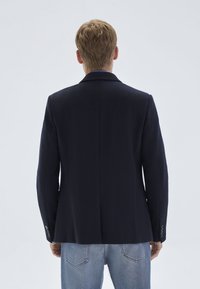 Person with short blond hair wearing a dark navy blazer and light blue jeans, standing facing away against a plain light background.