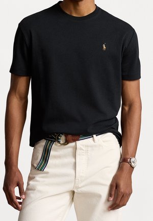 Mann in einem schwarzen Polohemd mit kurzen Ärmeln und einem kleinen gestickten Logo, beigen Hosen, gestreiftem Gürtel und einer braunen Lederuhr.