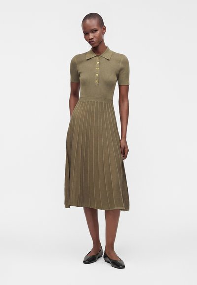 Femme portant une robe en maille côtelée vert olive à manches courtes, col, boutons dorés, jupe plissée et chaussures plates noires, debout sur fond blanc.