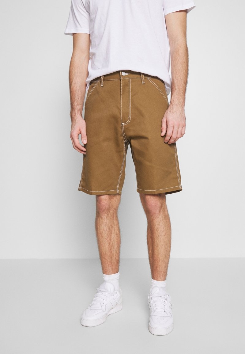 Shorts en coton marron avec des coutures blanches, coupe large, poches avant et fermeture à bouton, associés à des baskets blanches.