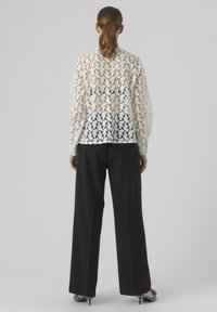 Blouse en dentelle à motif floral blanc, associée à un pantalon large noir. La blouse est dotée de poignets élastiques et d'une coupe décontractée.