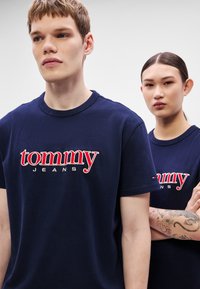 Dos personas llevan camisetas de algodón azul marino con texto bordado en blanco y rojo que dice "tommy JEANS". El tejido parece suave, con mangas cortas.
