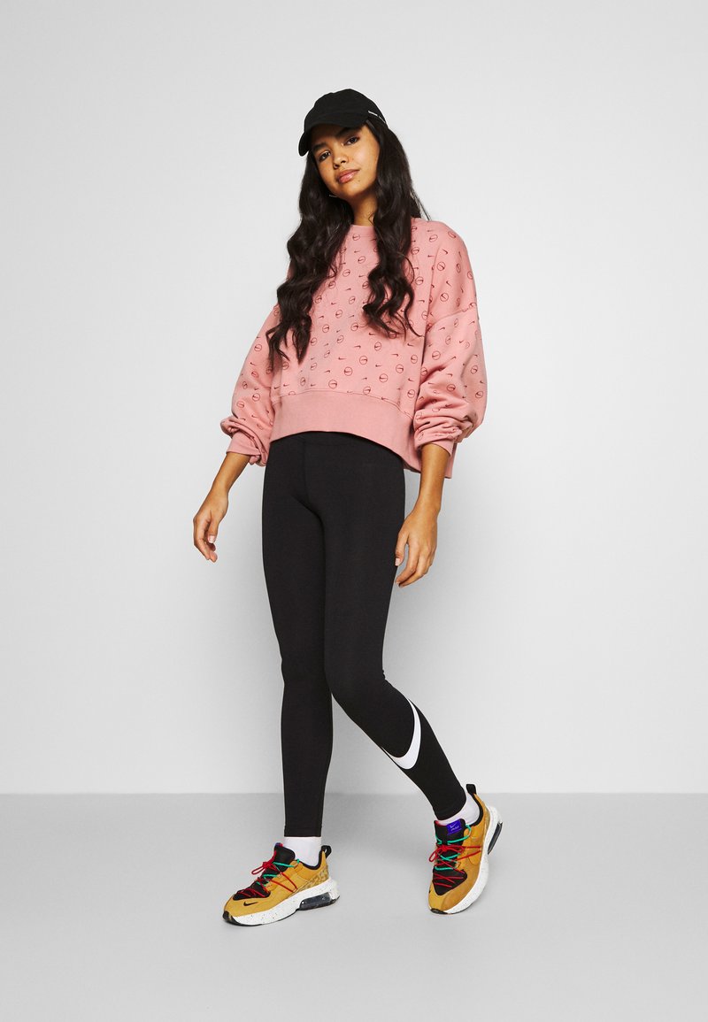 Sweatshirt rose avec des cercles texturés, leggings noirs et chaussures de sport multicolores avec un accent blanc et une casquette noire.