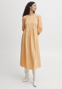 Een knielange jurk met pofmouwen in zachte perzikkleurige katoen, met een gathers in de taille en een gladde textuur, gecombineerd met witte hoge hakken.