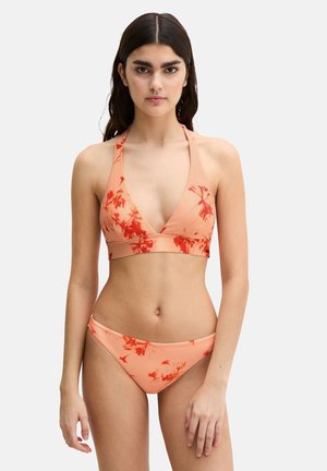 Kobieta ubrana w brzoskwiniowy bikini wiązany na szyi z czerwonymi kwiatowymi wzorami, stojąca na tle jednolitego białego tła, zwrócona przodem, z rękami swobodnie opuszczonymi.