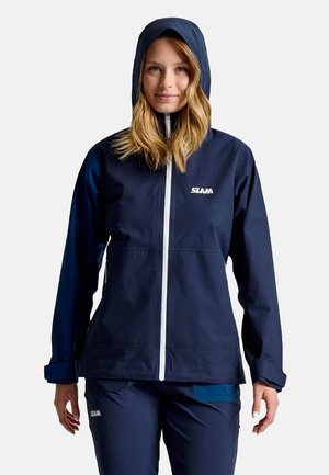 Veste imperméable marine avec un design ajusté, une capuche, une fermeture éclair blanche et un logo. Le matériau semble lisse et durable, adapté à une utilisation en extérieur.