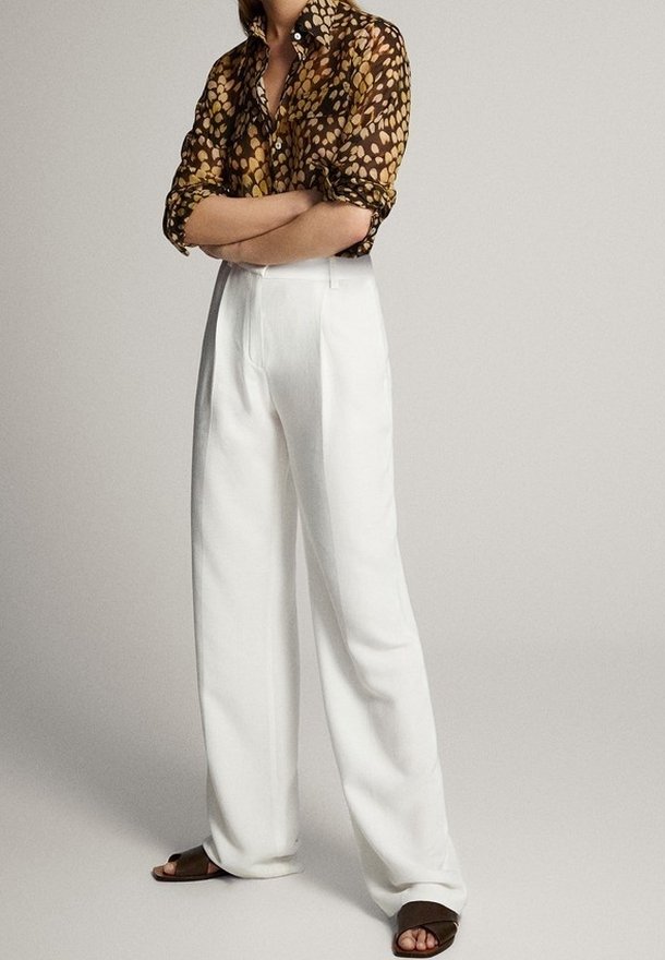 Personne portant une chemise marron à motifs avec les manches retroussées, un pantalon blanc taille haute à jambes larges et des sandales ouvertes marron foncé, debout les bras croisés.