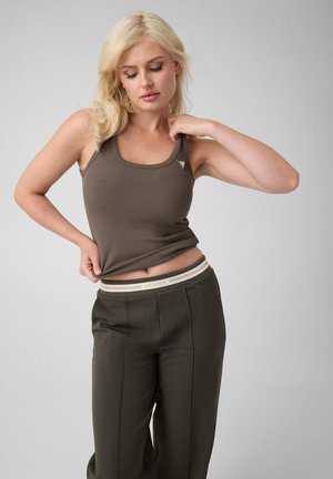 Femme blonde portant un débardeur olive et un pantalon ajuste son haut tout en regardant vers le bas sur un fond gris uni.